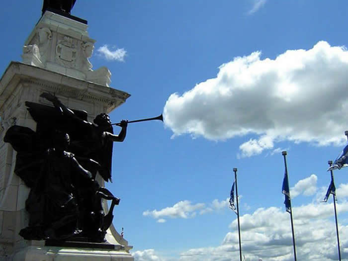 A perfectly timed forced-perspective photo of a dark bronze statue playing a trumpet. The end of the trumpet is positioned against a bright white cloud in a blue sky, making it look like the cloud is smoke or sound coming from the instrument.