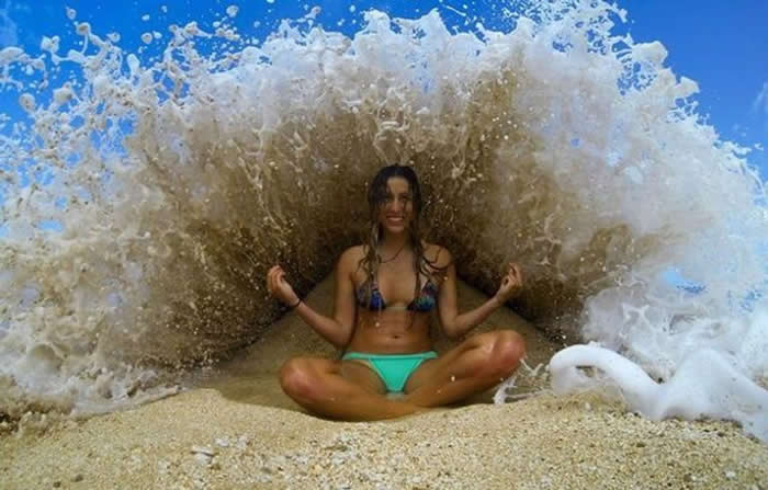 A perfectly timed action shot of a woman sitting cross-legged on a sandy beach. A large ocean wave is bursting upward directly behind her, creating a symmetrical arc of white foam and spray that looks like a liquid throne.