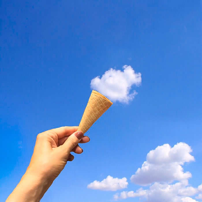 A person&rsquo;s hand holds an empty waffle ice cream cone up toward a clear blue sky. A single, white, puffy cloud is positioned exactly at the top of the cone, creating the illusion of a scoop of fluffy ice cream.