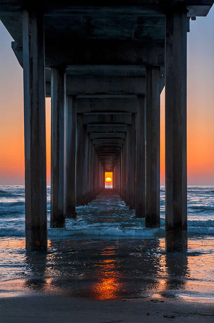 A symmetrical, low-angle shot from beneath a concrete pier extending into the ocean. The sun is setting exactly in the center of the far opening, framed by two rows of dark pillars, reflecting golden light onto the wet sand.