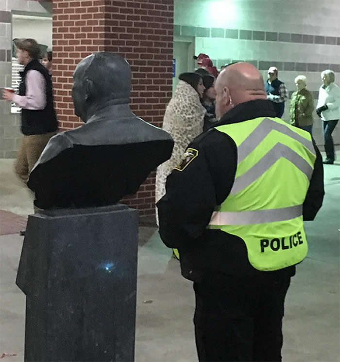 A funny candid photo from behind a bald police officer wearing a neon yellow "POLICE" vest. He is standing next to a bronze statue of a man's head and shoulders, whose bald head perfectly matches the officer's in shape and size.