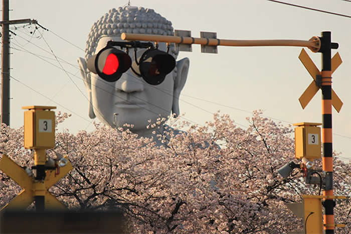 A forced-perspective photo of a giant gray Buddha statue behind a railway crossing. The two circular traffic signal lights are perfectly aligned with the statue's eyes, making it look like the Buddha is wearing large, dark goggles with one red light glowing.