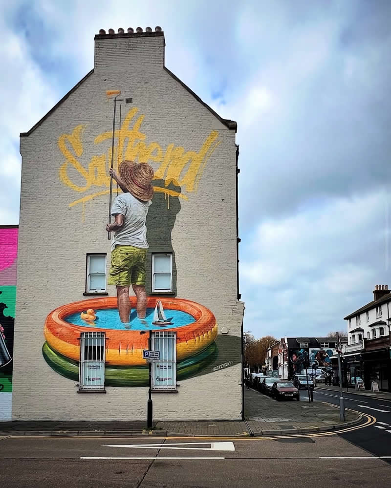 A large mural on the side of a building shows a man standing in an inflatable kiddie pool, painting the word &ldquo;Stay Fresh&rdquo; in bright yellow across the wall, creating a playful illusion in an urban street setting.