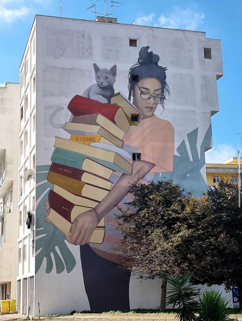 A large mural on a building shows a young woman with glasses holding a tall stack of books, with a gray cat perched on top. The artwork blends soft colors and urban surroundings, with trees partially covering the lower section.