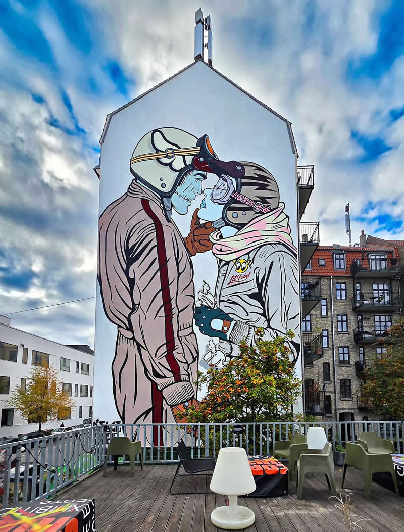 A large mural on the side of a building depicts two people wearing vintage motorcycle helmets leaning in for a kiss. The artwork stands against a cloudy sky, with a terrace and chairs in the foreground and residential buildings nearby.
