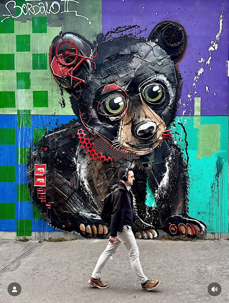A large street mural depicts a bear made of mechanical and recycled parts with big expressive eyes, set against a colorful geometric background. A man wearing headphones walks past the artwork in the foreground.