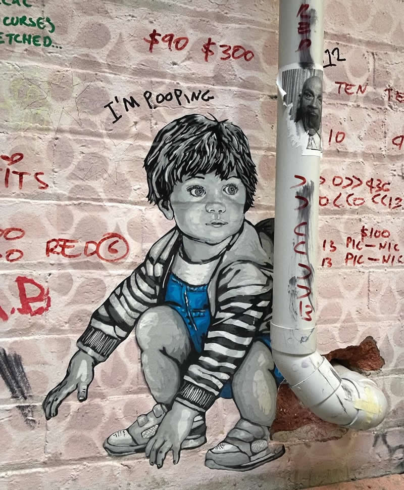 A street artwork on a brick wall shows a young child squatting, humorously interacting with a real drainpipe positioned behind him, with graffiti text and markings surrounding the scene.