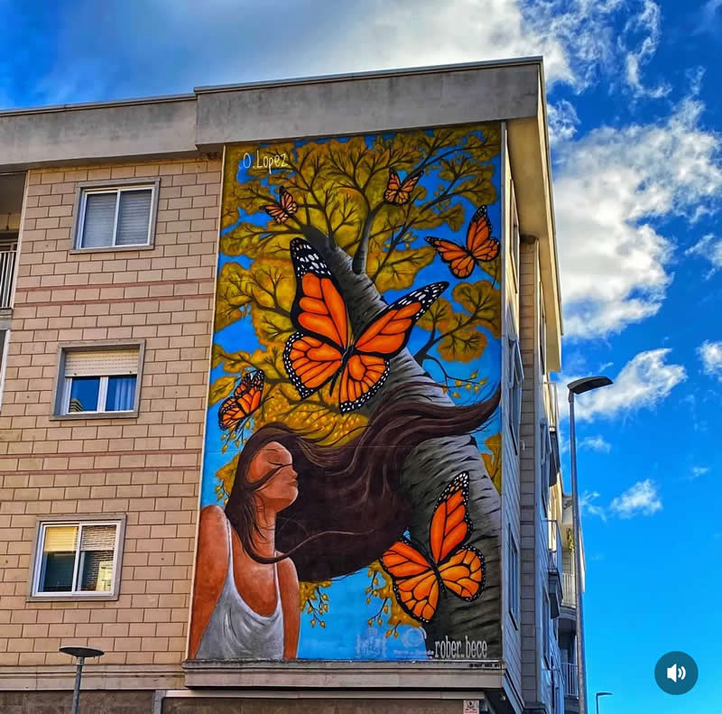 A colorful mural on a building shows a woman with flowing hair blending into a tree, surrounded by vibrant orange butterflies against a bright blue sky, symbolizing transformation and harmony with nature.