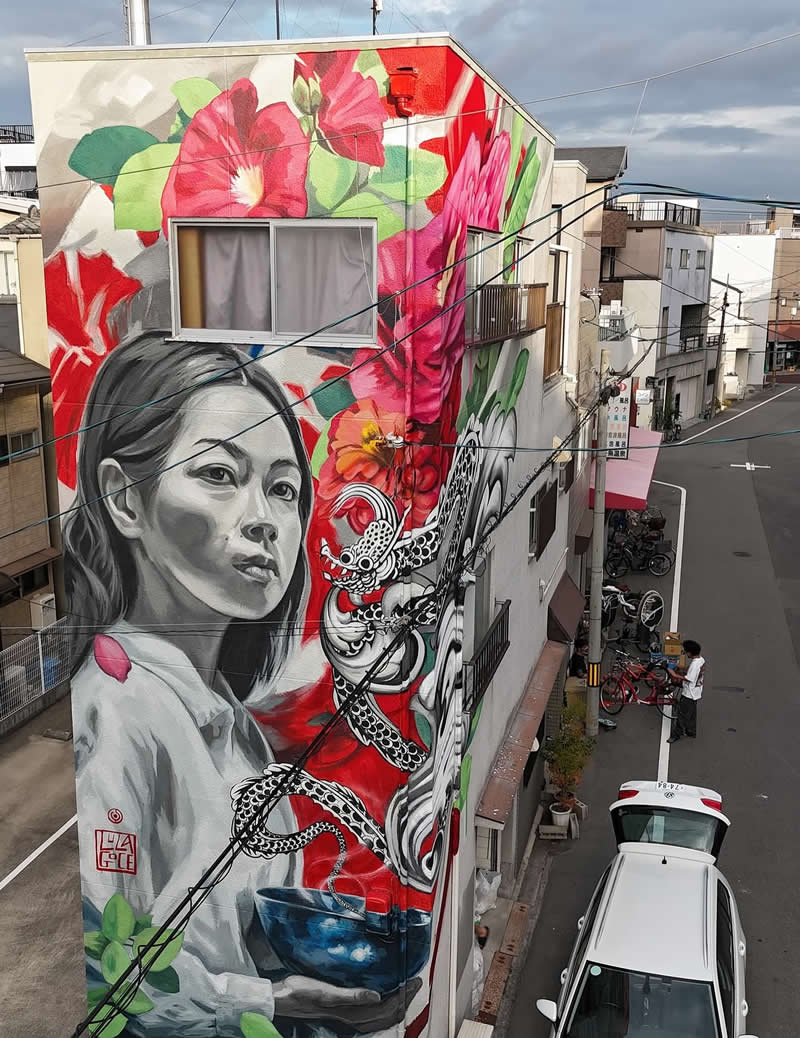 A large mural on a building features a grayscale portrait of a woman holding a bowl, surrounded by vivid red and pink flowers and a detailed dragon, creating a striking contrast against the urban street below with cars and power lines.