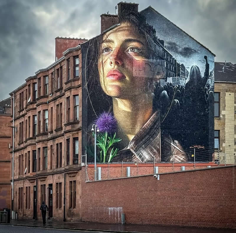 A large mural on the side of a building shows a realistic portrait of a young woman gazing upward, blended with scenes of a crowd protest and a blooming flower, symbolizing resilience and social change in an urban setting.