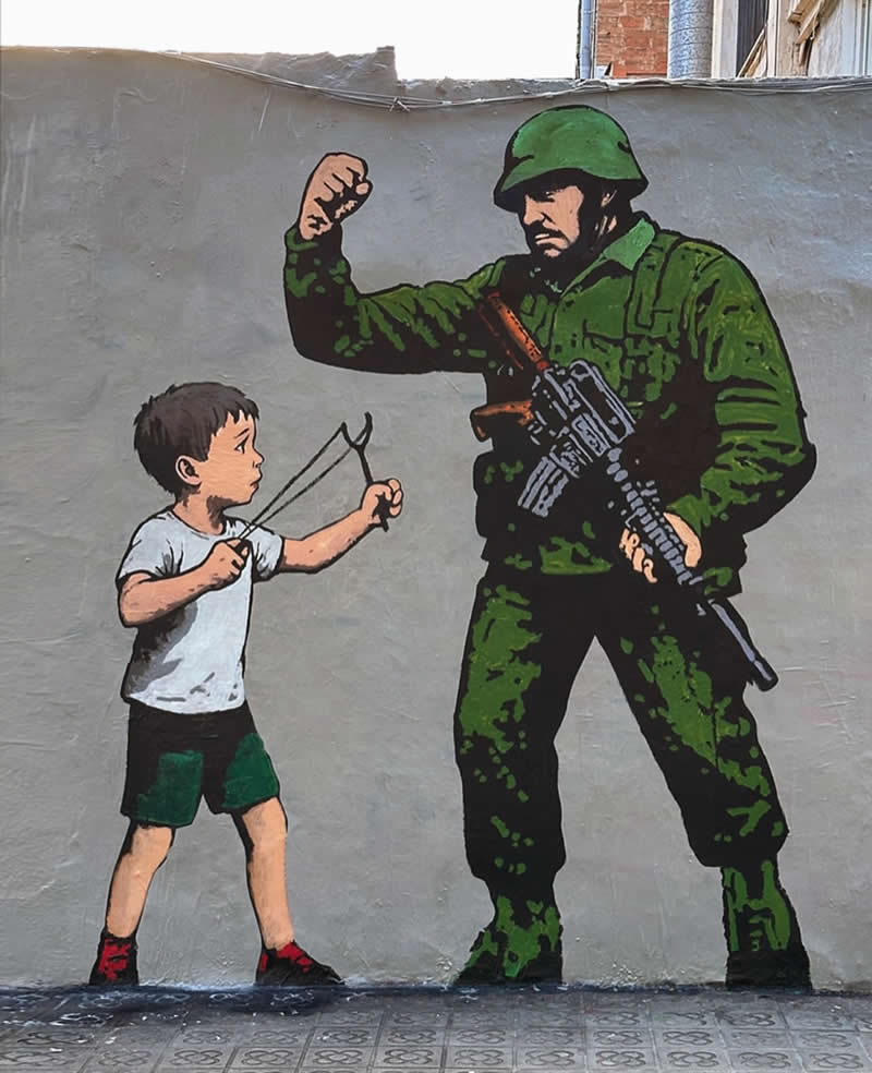 A street mural shows a young boy aiming a slingshot at an armed soldier in green uniform, highlighting a stark contrast between innocence and military force on a plain wall background.