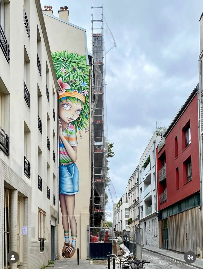 A tall mural on the side of a narrow building shows a stylized girl with leafy green hair and flowers, wearing a colorful outfit, blending nature with city architecture along a quiet street.