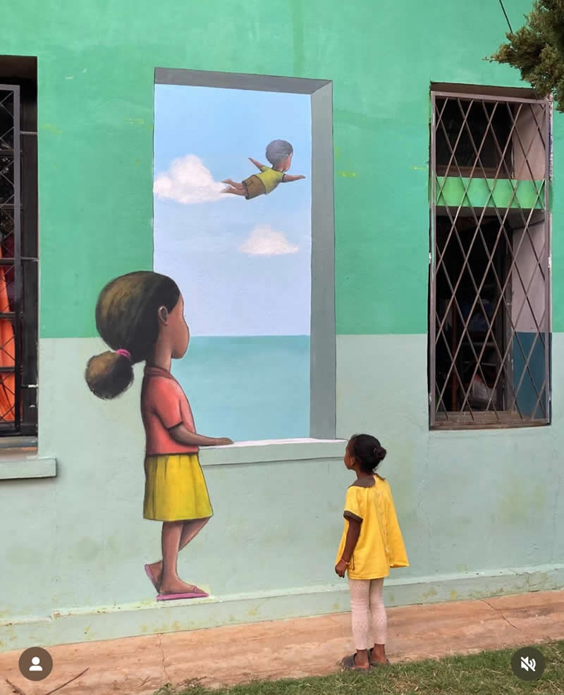 A mural on a green wall shows a painted girl looking out of a window at a flying child in the sky, while a real child stands in front of the artwork, blending reality with imagination.