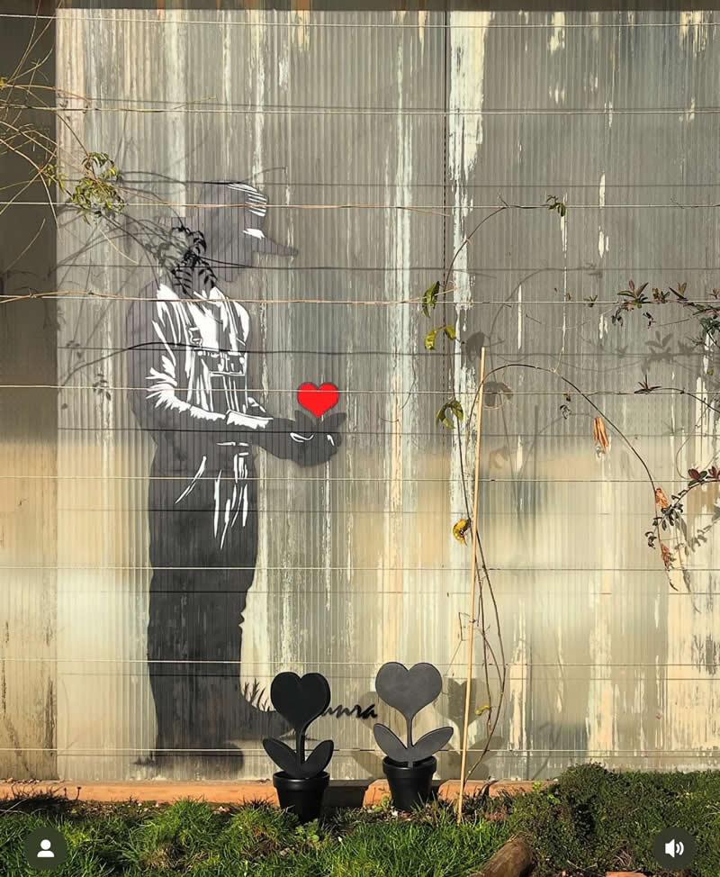 A stencil-style street artwork shows a figure in monochrome holding out a bright red heart, painted on a weathered wall with plants and small heart-shaped decorations placed below.