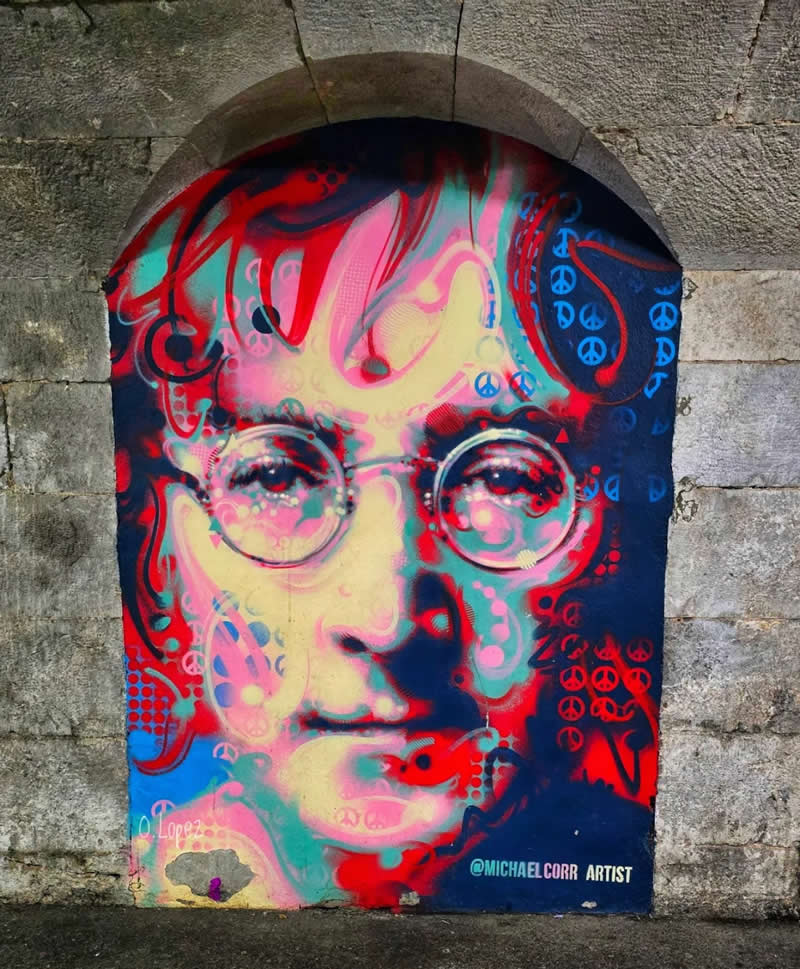 A vibrant street mural inside an arched stone wall features a colorful, abstract portrait of a man with round glasses, painted in bold red, blue, and neon tones with swirling patterns and peace symbols.