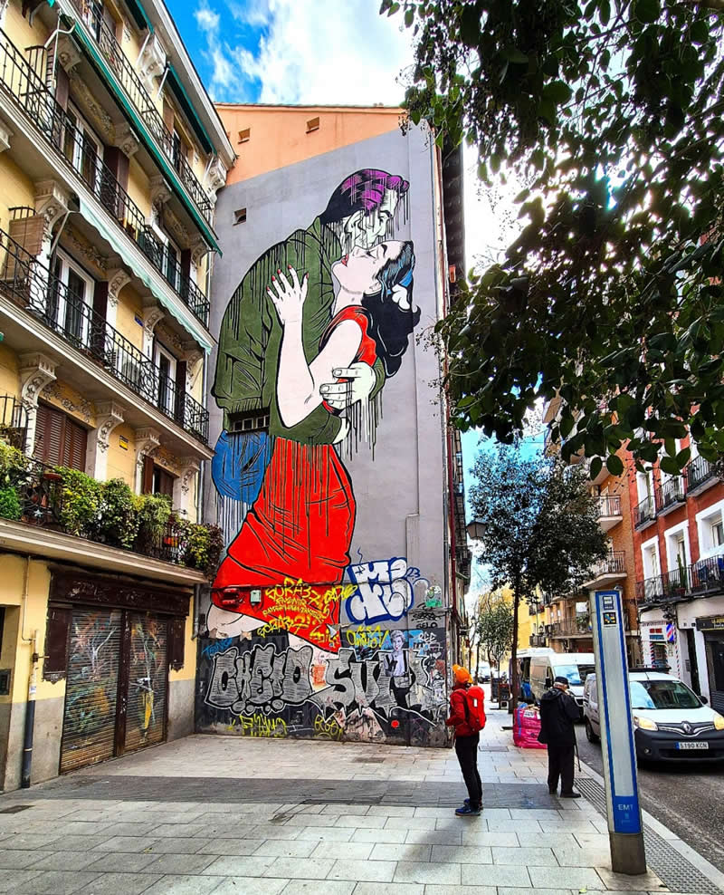 A large mural on a city building shows a couple embracing and kissing, with dripping paint effects and bold colors, surrounded by urban streets, balconies, and pedestrians below.
