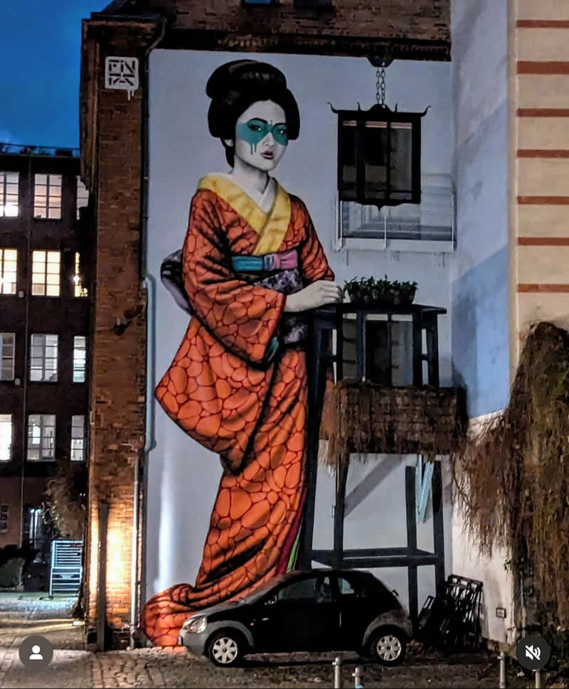 A large mural on a building depicts a geisha in a detailed orange kimono with stylized makeup, towering above a parked car in a dimly lit urban alley, blending traditional Japanese aesthetics with a contemporary city setting.