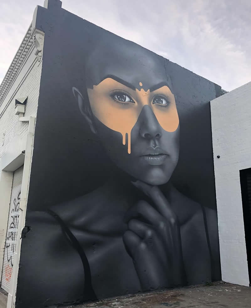 A large monochrome mural of a woman&rsquo;s face with a thoughtful expression, partially covered by a dripping golden mask around her eyes, painted on a dark wall in an urban setting.