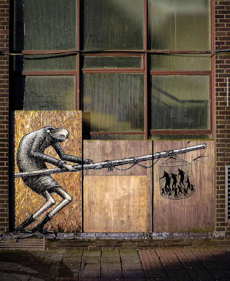 A street artwork on wooden panels shows a surreal figure pulling a long pole attached to a cage filled with silhouetted human figures, symbolizing control and manipulation against an industrial building backdrop.