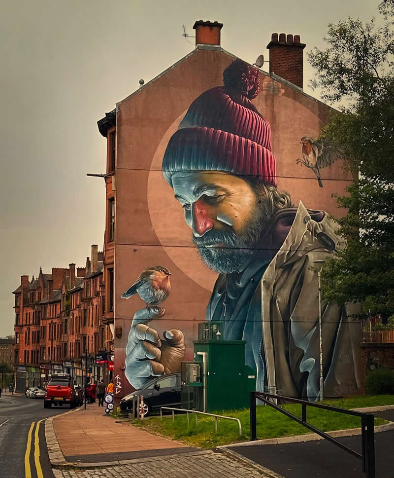 A large mural on a building shows a bearded man wearing a knit hat gently holding a small bird on his finger, with another bird nearby, set against a warm-toned urban street backdrop.