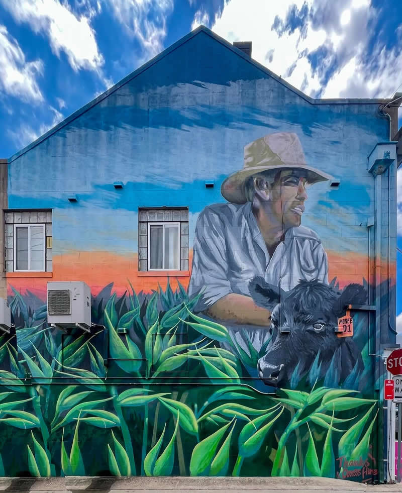 A large mural on a building depicts a farmer wearing a hat leaning over lush green crops, with a cow beside him, set against a colorful sky backdrop in an urban street setting.