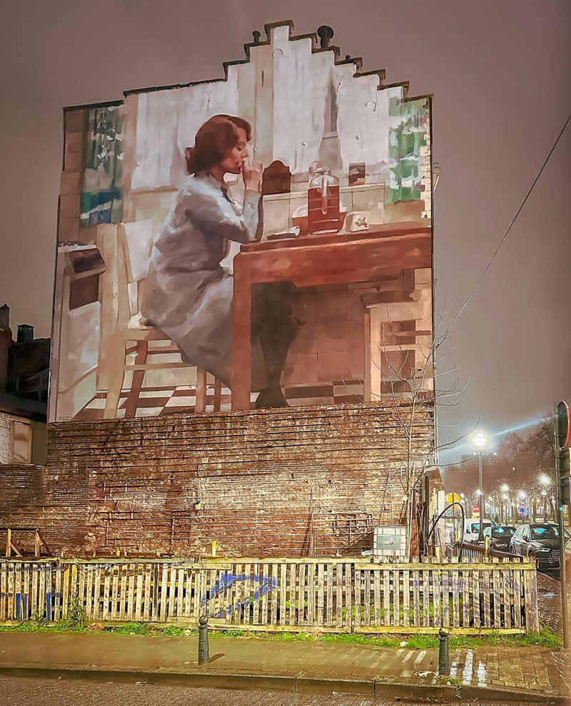 A large mural on a building depicts a woman sitting at a kitchen table, holding a cup in a softly lit interior scene, contrasting with the nighttime urban street outside.