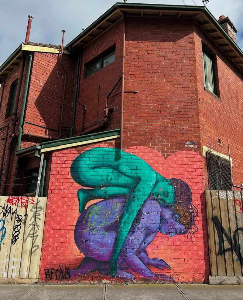 A street mural on a brick wall shows two intertwined human figures in contrasting colors, one crouched while the other leans heavily on top, symbolizing emotional weight and support in an urban alley setting.