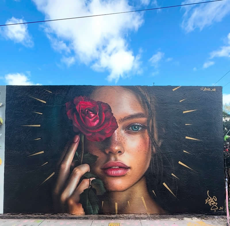 A hyper-realistic mural shows a young woman&rsquo;s face with one eye covered by a red rose, surrounded by subtle golden rays on a dark background under a bright blue sky.