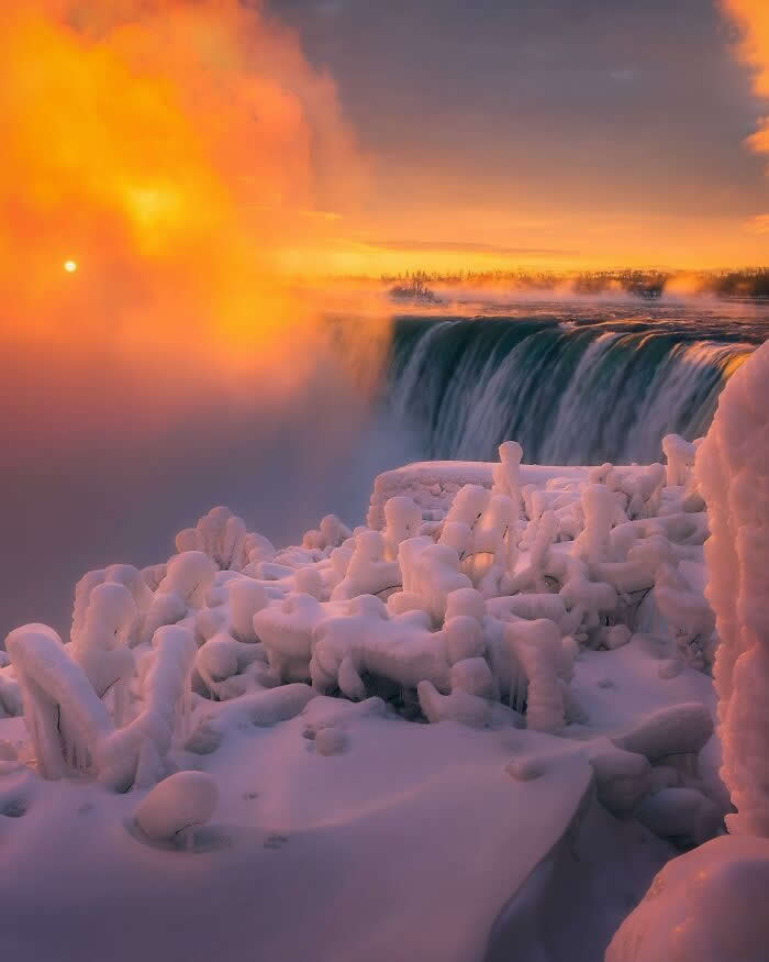 Golden sunrise light glows through icy mist over frozen Niagara Falls, with snow-covered formations and dramatic winter scenery surrounding the cascading waterfall.