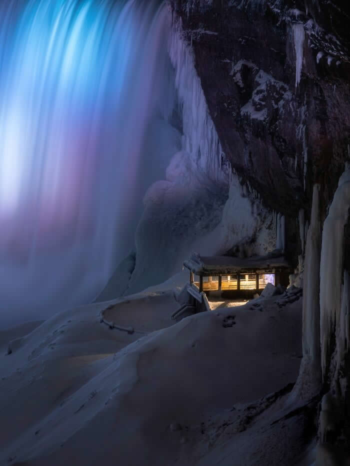 A softly lit shelter sits beneath towering frozen waterfalls and icy cliffs at night, surrounded by snow and glowing blue mist in a dramatic winter landscape.