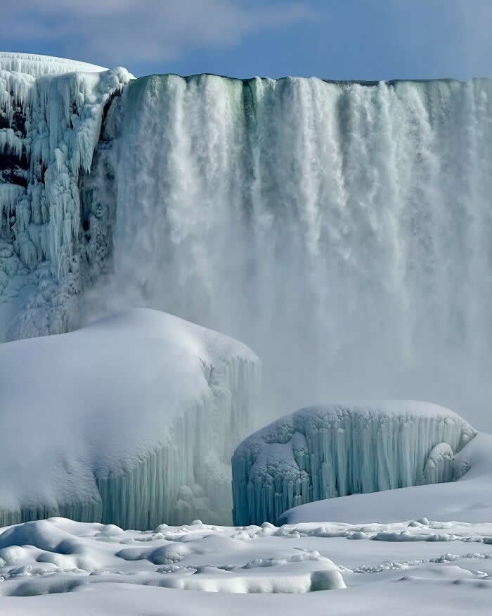 Massive icy waterfalls cascade over Niagara Falls in winter, surrounded by snow-covered formations and frozen cliffs beneath a cold blue sky.