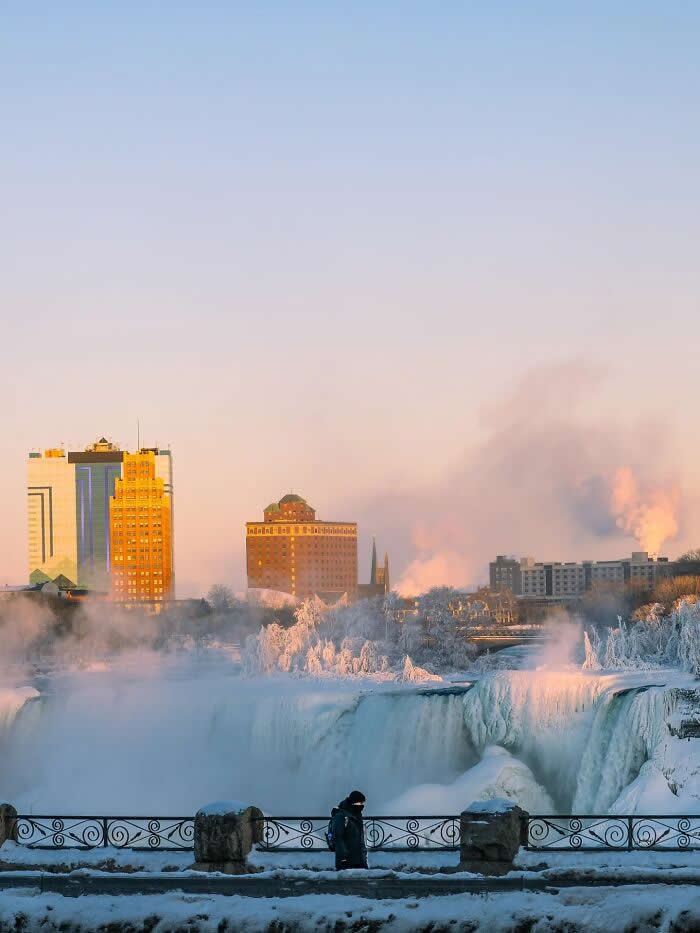 A lone person walks along a snowy railing overlooking frozen Niagara Falls at sunrise, with mist rising around icy waterfalls and city buildings glowing in warm morning light.