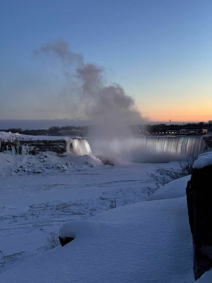 Mist rises above partially frozen Niagara Falls at dusk, surrounded by deep snow and icy cliffs beneath a soft blue and orange evening sky.