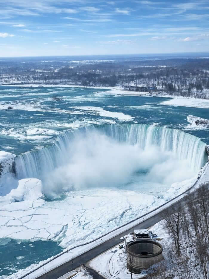 Aerial view of Niagara Falls surrounded by snow and frozen river ice, with powerful waterfalls plunging into a mist-filled basin beneath a bright winter sky.