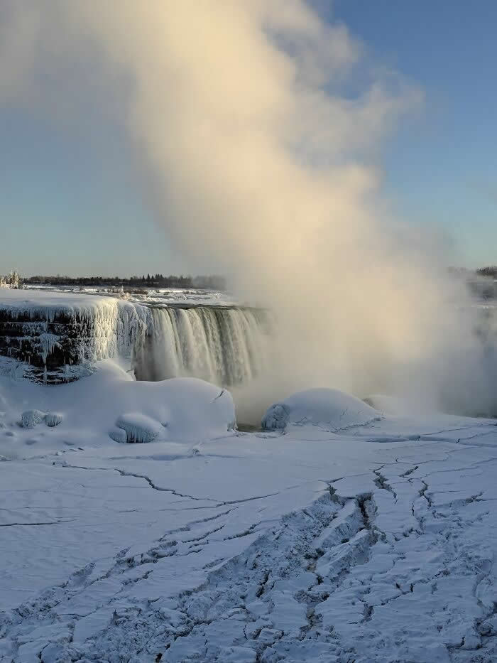 Towering mist rises above frozen Niagara Falls surrounded by snow-covered ice formations and cracked icy ground under a clear winter sky.