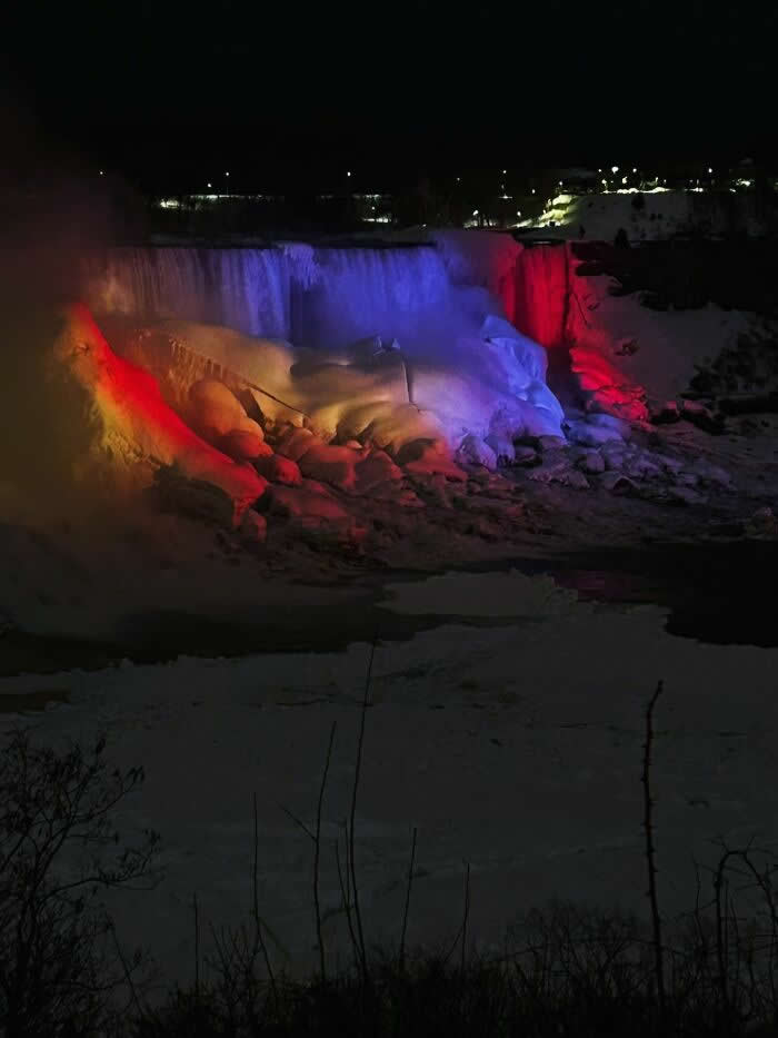 Niagara Falls glows with dramatic red and blue lights at night, illuminating frozen rocks, icy waterfalls, and snowy winter scenery beneath a dark sky.