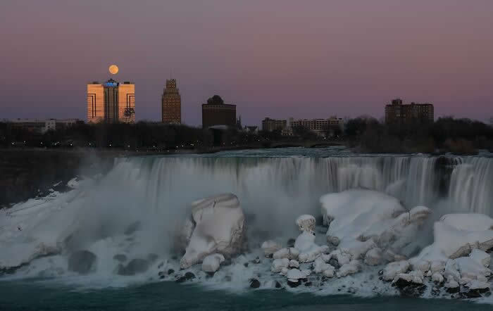 The moon rises above city buildings overlooking frozen Niagara Falls at dusk, with icy rocks and cascading waterfalls glowing beneath a soft purple evening sky.