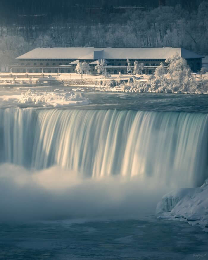 Smooth cascading waters flow over Niagara Falls beside snow-covered trees and buildings, creating a serene winter scene with soft mist and icy surroundings.