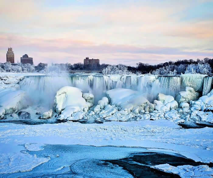Snow-covered Niagara Falls cascades through icy cliffs and frozen riverbanks, with soft pastel skies and distant city buildings creating a peaceful winter panorama.