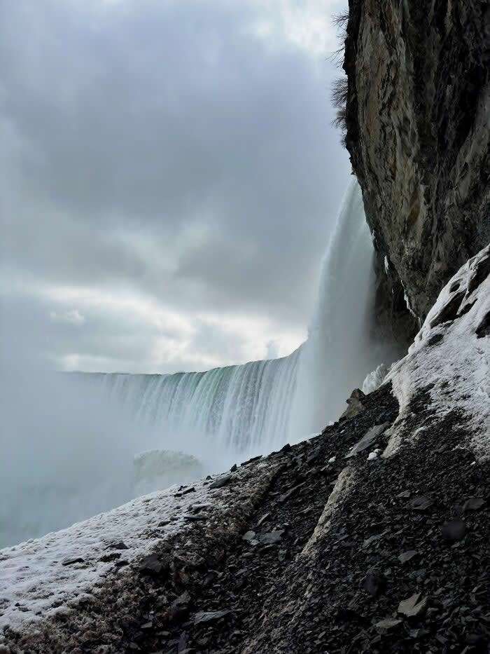 A dramatic side view of Niagara Falls cascades beside rocky snow-covered cliffs, with mist rising beneath dark cloudy skies in a rugged winter landscape.