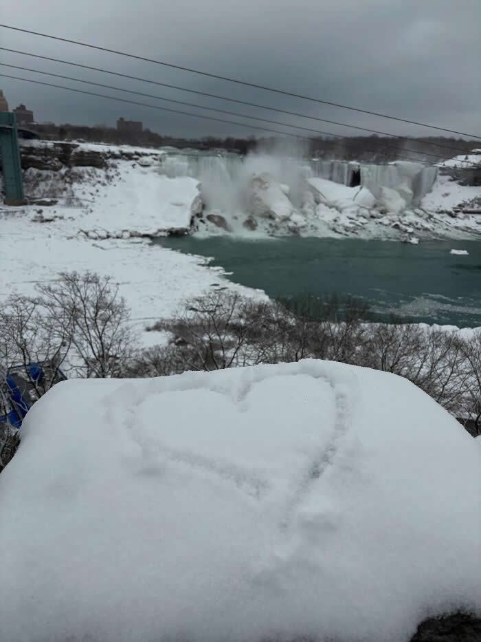 A heart shape drawn in fresh snow overlooks icy Niagara Falls and frozen riverbanks beneath a gray winter sky.