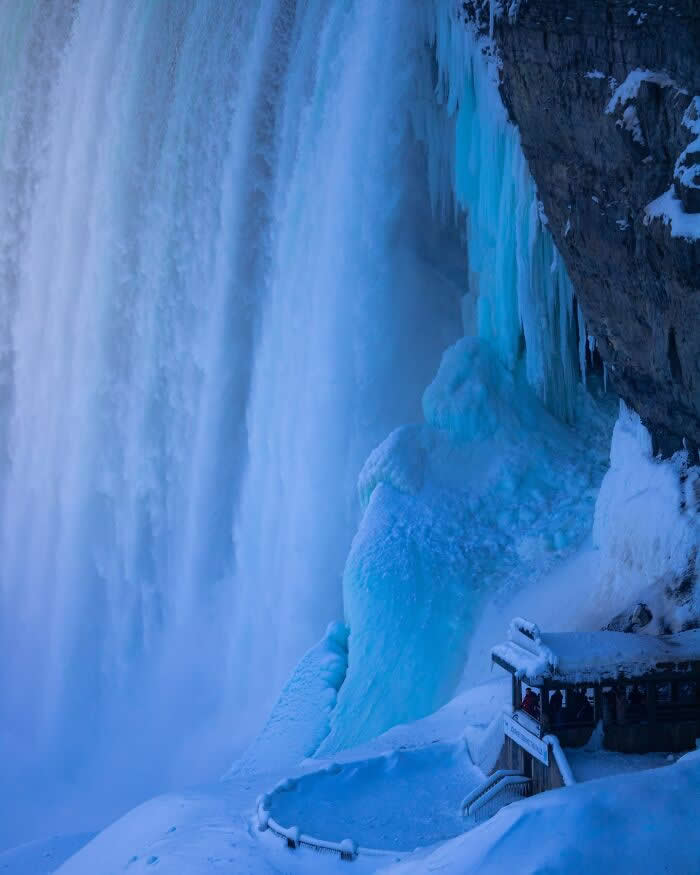 Towering frozen waterfalls and blue ice formations surround a snow-covered viewing platform beneath Niagara Falls in a dramatic winter scene.