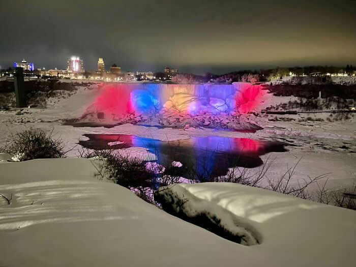 Niagara Falls glows with pink and blue lights at night, reflecting across icy water and snow-covered landscapes with city lights shining in the background.