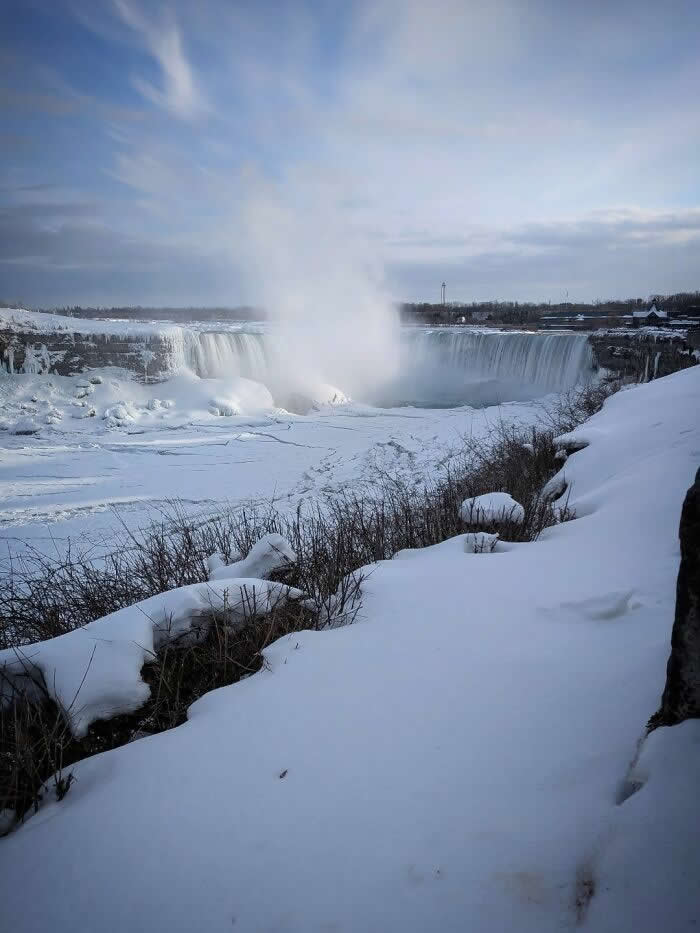 Snow-covered cliffs and frozen terrain surround Niagara Falls as towering mist rises into a pale blue winter sky above icy waterfalls.