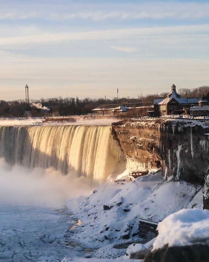 Warm sunlight illuminates Niagara Falls as icy cliffs, snow-covered paths, and frozen mist create a glowing winter landscape beside the cascading waterfall.