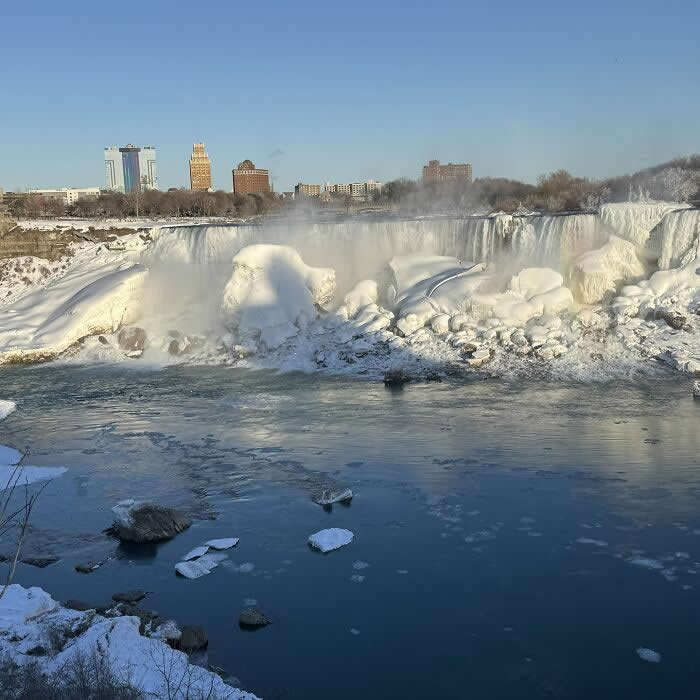 Frozen Niagara Falls rises above icy blue water and snow-covered rock formations, with city buildings visible beneath a clear winter sky.