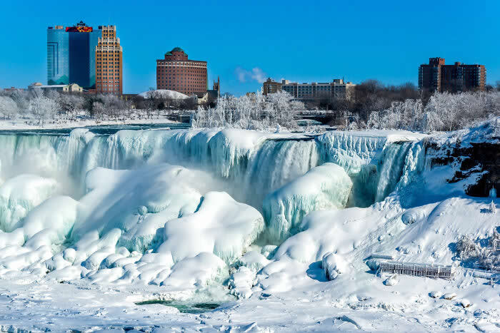 Snow and ice blanket Niagara Falls beneath a vivid blue sky, with frozen waterfalls, icy formations, and distant city buildings creating a striking winter panorama.