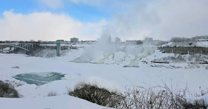 A wide snowy landscape surrounds frozen Niagara Falls as thick mist rises above icy waters and distant bridges beneath a pale winter sky.