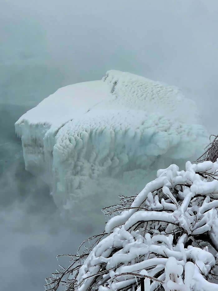 A massive ice-covered formation rises through thick mist near Niagara Falls, with snow-covered branches framing the quiet winter scene.