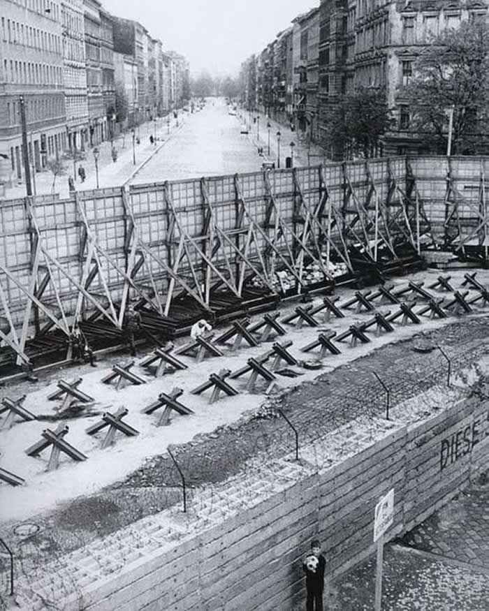 A boy plays in the shadow of the Berlin Wall while soldiers stand guard on the other side. Berlin Wall, Bernauer Strasse, West Berlin 1967.